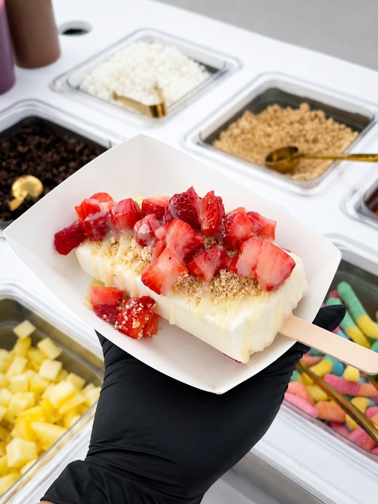 A milk-based paleta topped with fresh strawberries and graham cracker crumbs, held over the full topping bar
